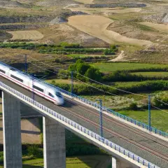 Un tren de alta velocidad cruza un viaducto sobre un paisaje rural. El tren, que parece ir de Lleida a Madrid, es plateado con una franja morada.