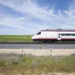 Un moderno tren blanco con detalles morados avanza por las vías. El paisaje verde y el cielo azul sugieren un viaje rápido de A Coruña a Madrid.