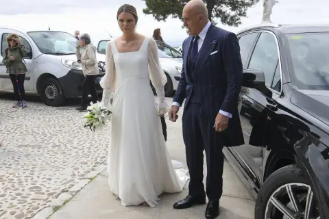 Novia con ramo de lirios y su acompañante, un hombre con traje de raya diplomática, llegan a su boda. Irene Matamoros, radiante.
