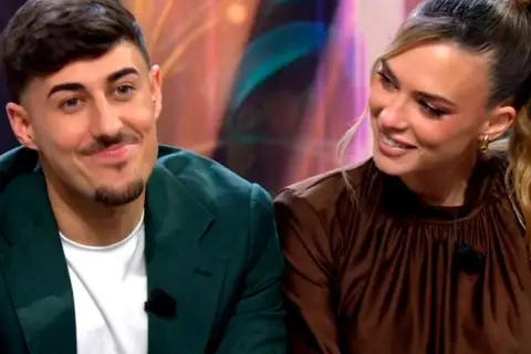 Almudena y Borja, sonriendo y mirando hacia un lado. Él lleva una chaqueta verde y ella un vestido marrón.