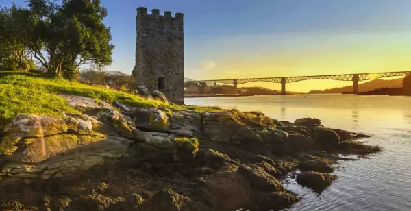 Torre medieval junto a un río al atardecer, con un puente al fondo. Imagina el tren Madrid Ferrol pasando por allí.